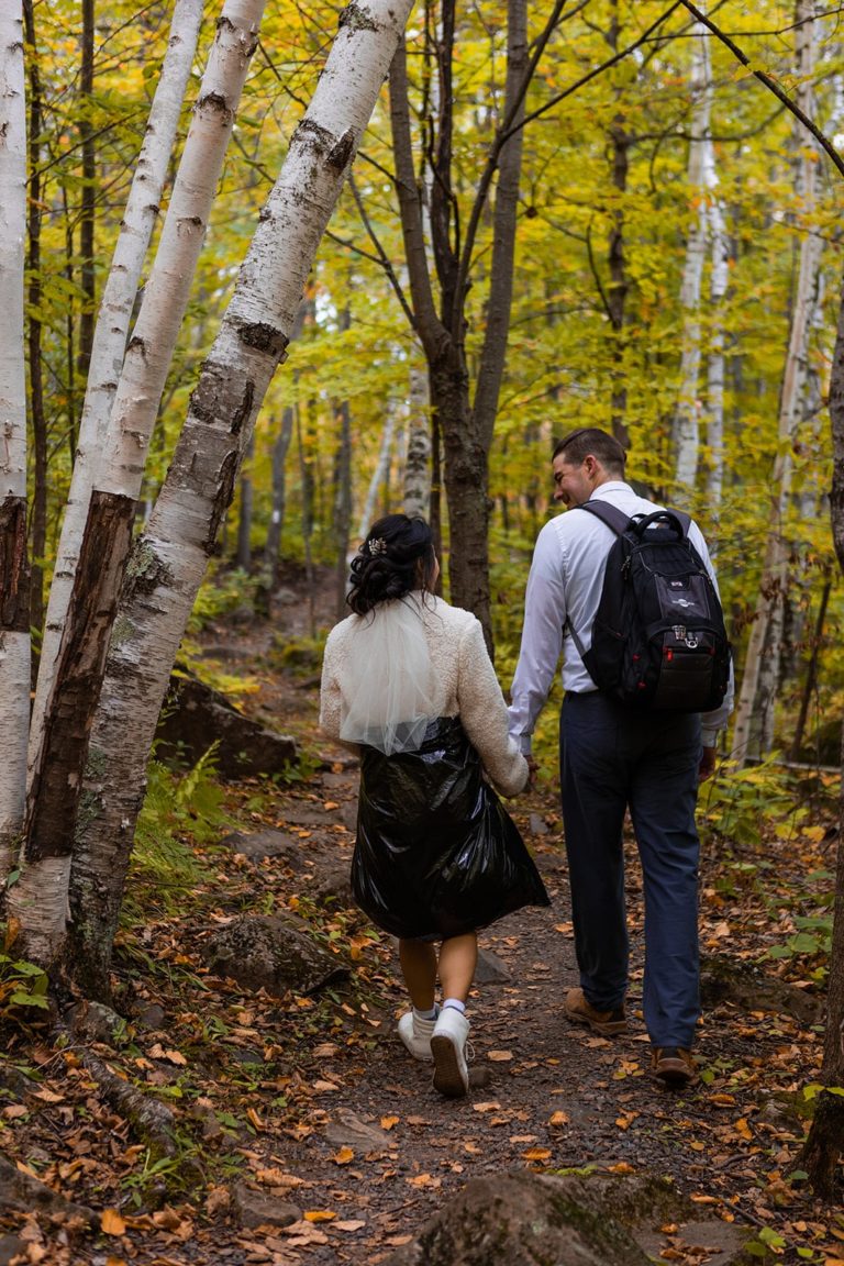 A bride and groom smile at one another as they hike through a forest of fall leaves during their fall adventure elopement along the North Shore of Lake Superior in Duluth, MN. Hiking in their wedding clothes during their Duluth elopement was part of the excitement of the day for this adventure loving couple.