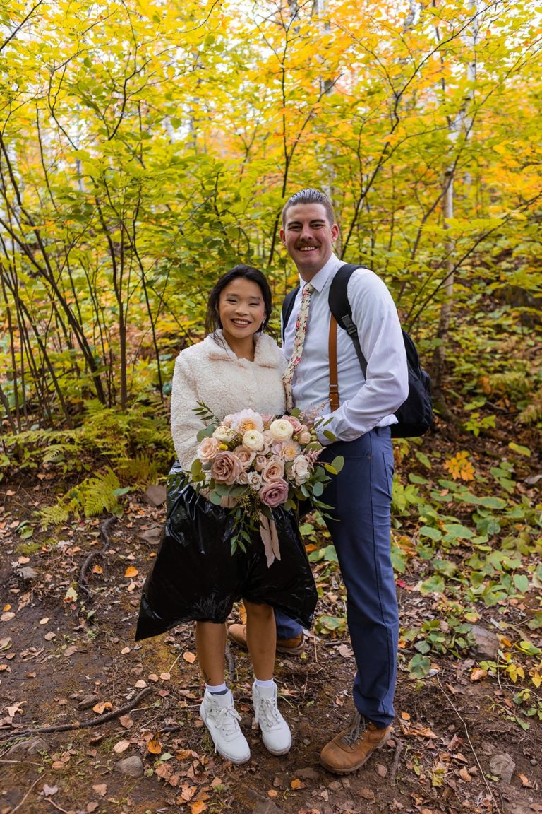 A bride and groom smiles at the camera among a forest of fall leaves. The bride is wearing a cute white jacket and a black trash bag. Her wedding dress is tucked into the trash bag to keep it clean while she hike in wedding dress during her fall adventure elopement along the North Shore of Lake Superior in Duluth, MN.