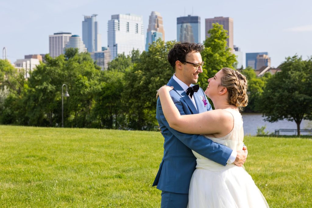 A couple embrace in a grassy field on a beautiful summer day in front of the Minneapolis skyline. Summer is one of the best times of year to get married in Minnesota.
