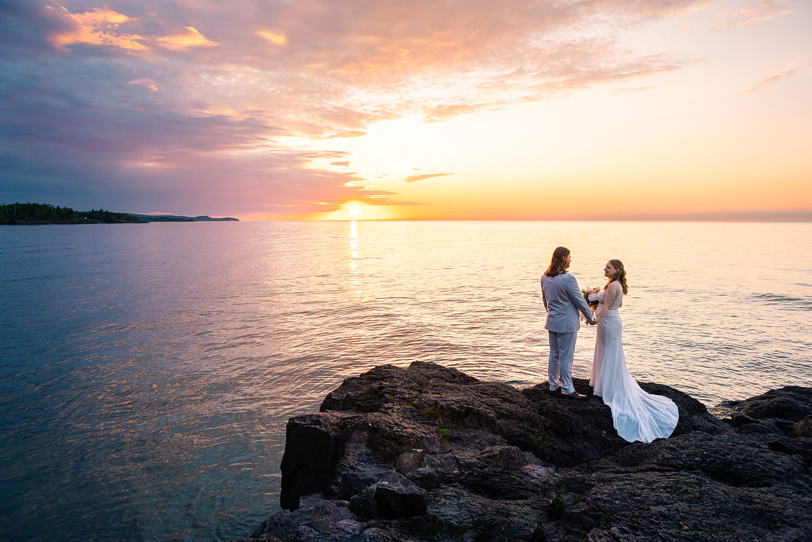A couple watch the sunrise over Lake Superior during their North Shore elopement in Minnesota. The sun glows golden over the water as they watch from a rocky outcropping on the shore.