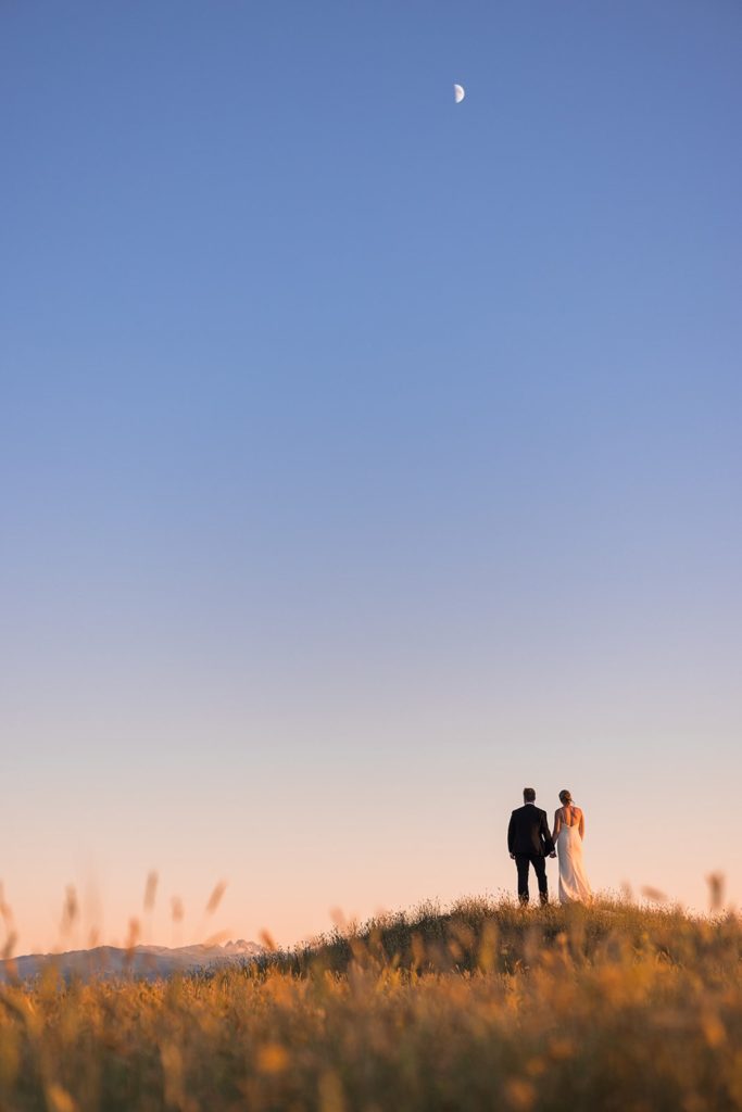 A bride and groom stand on a grassy hill in a golden field looking off into the distance at the Bighorn Mountains and a crescent moon rising in the sky as the sunsets.