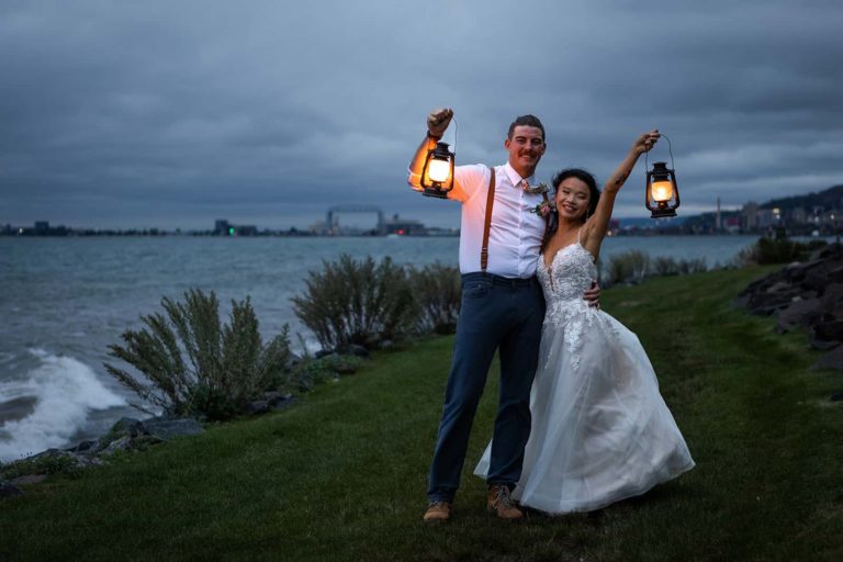 A bride and groom smile at the camera on the moody shore of Lake Superior during their elopement in Duluth, MN. The weather is overcast and they hold lanterns that glow brightly and light up their faces.