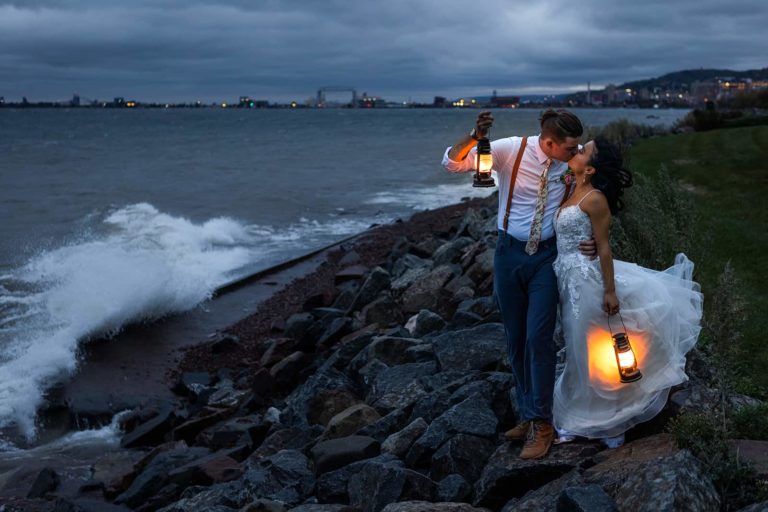 A bride and groom kiss on the moody shore of Lake Superior during their elopement in Duluth, MN. The weather is overcast and they hold lanterns that glow brightly.