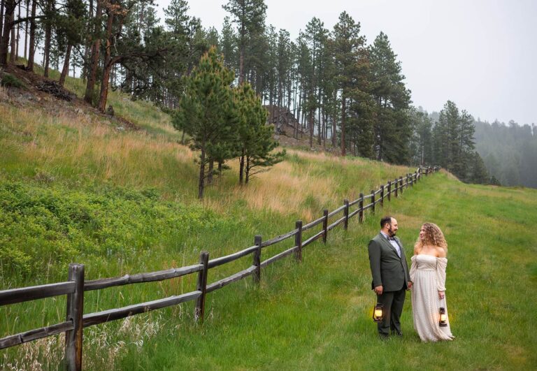 A couple in their wedding clothes walks along an old wooden fence during their Black Hills elopement in South Dakota. It's an overcast day and they hold lanterns to light their way.