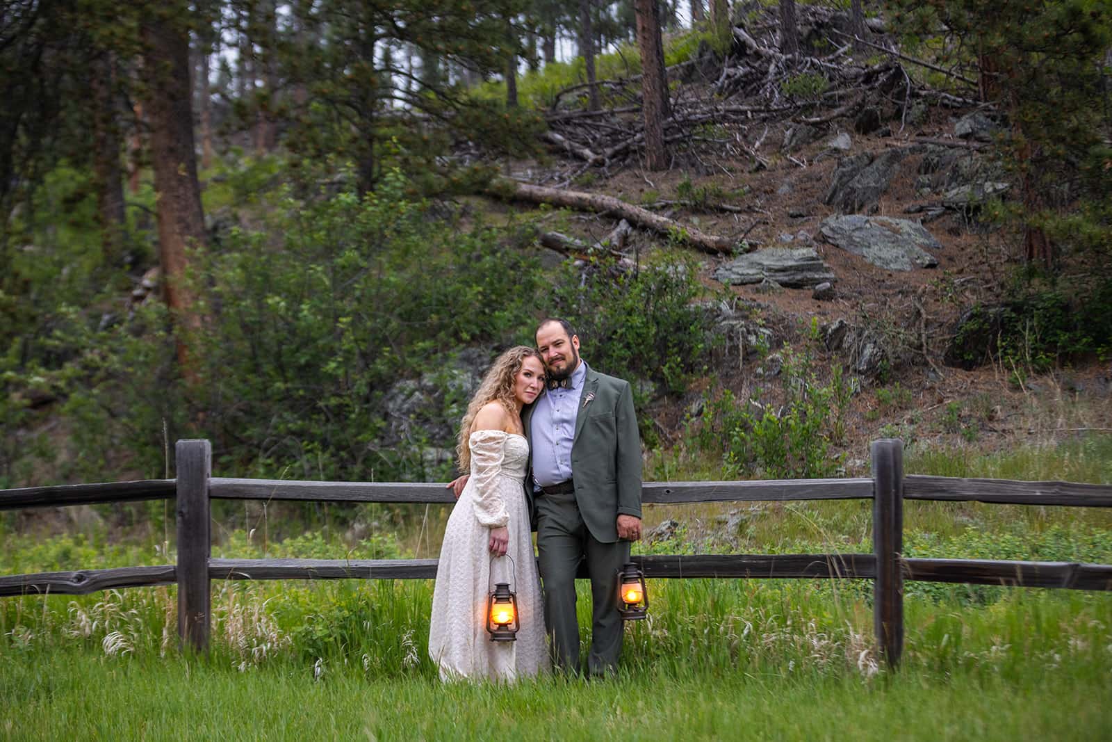 A couple stands in front of an old wooden fence holding glowing lanterns during their Black Hills elopement in South Dakota. Behind them, towering pines grow of a rocky hill. The backdrop is a perfect example of the beauty that the Black Hills of South Dakota.