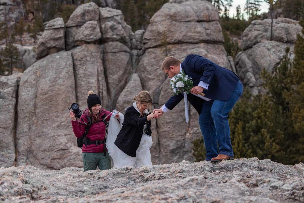 Black Hills South Dakota elopement photographer Forever and Evergreens' Tali helps a bride scramble up a rocky path during an adventure elopement in Custer State Park near Hill City, SD.