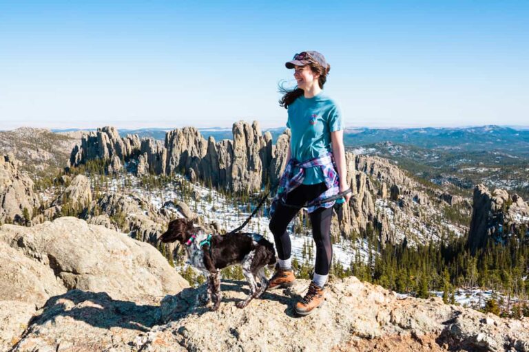 A woman and her dog smile off into the distance as the wind whips her hair around. Behind them, the unmistakable rock formations of the Black Hills jut into the sky.