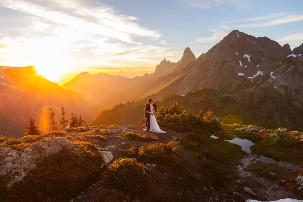 Standing in the golden glow of sunset, a bride and groom stare into one another's eyes during their Mount Baker elopement. Around them, the mountains of the North Cascades glow with the hazy light of sunset.