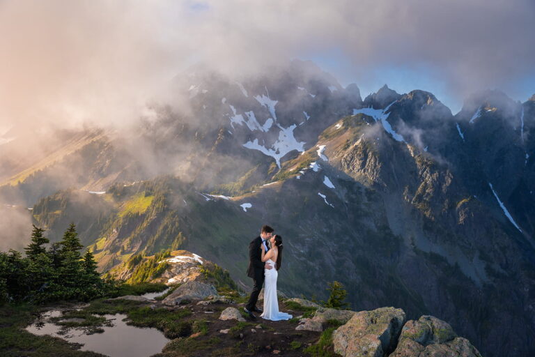 A couple share a kiss during their Mount Baker elopement in front of a large mountain with clouds rolling in front.