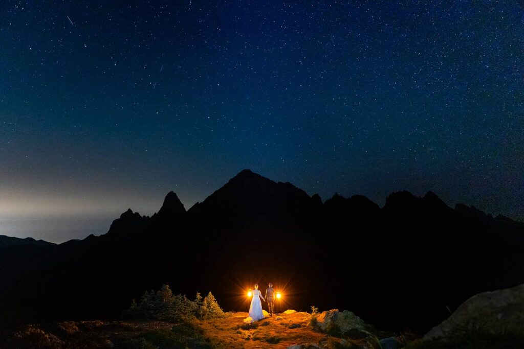 A couple hold lanterns and stand in front of a large mountain under the stars during their Mount Baker elopement in Washington State.