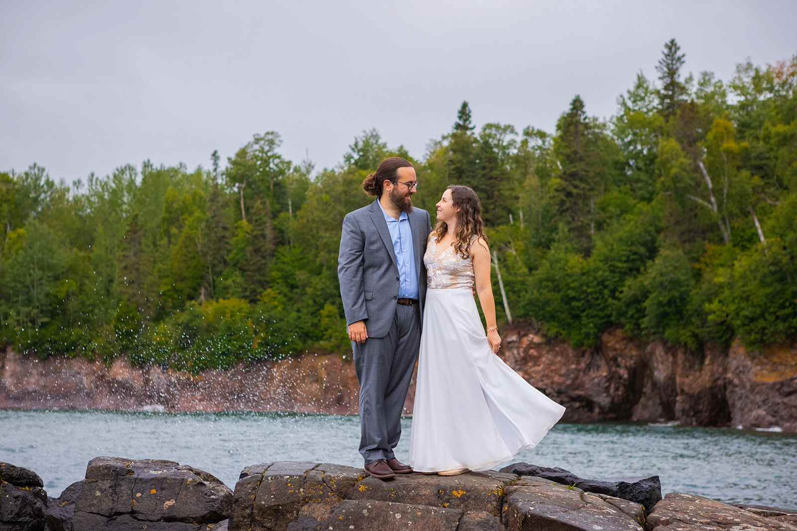 Waves crash along the shore behind a couple smiling at one another in their wedding clothes in Tettegouche State Park.