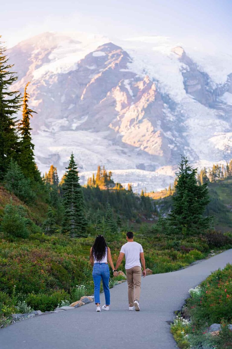 A man and woman hold hands and walk down a paved path in front of a large snow capped mountain.