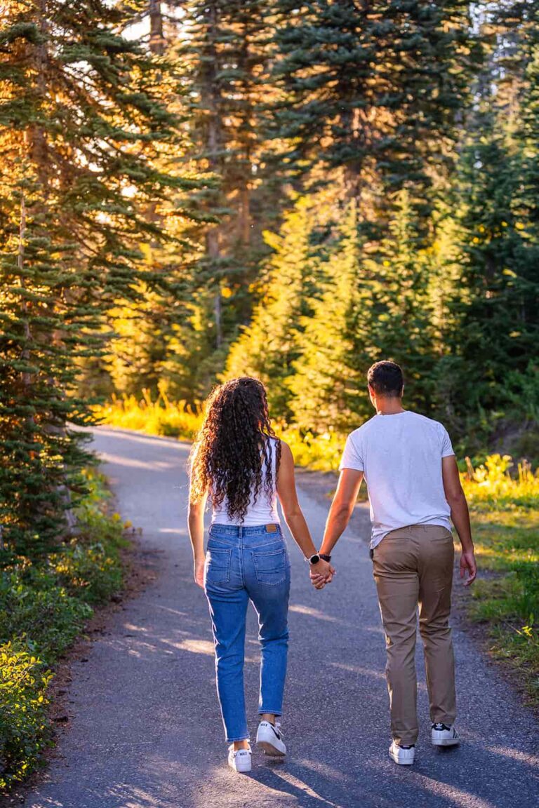 A couple holds hands as they walk along a paved trail through a pine forest in Washington state.
