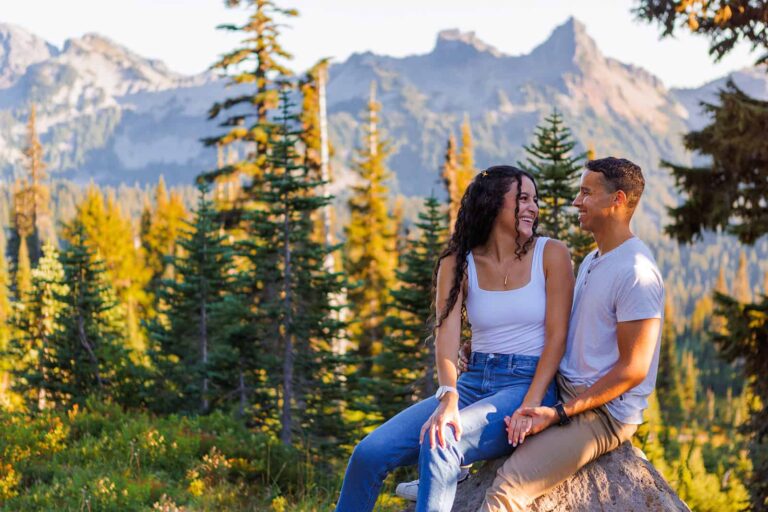 A couple sits on a rock in front of a mountain ridge line and a pine forest in Washington state during their engagement photos.