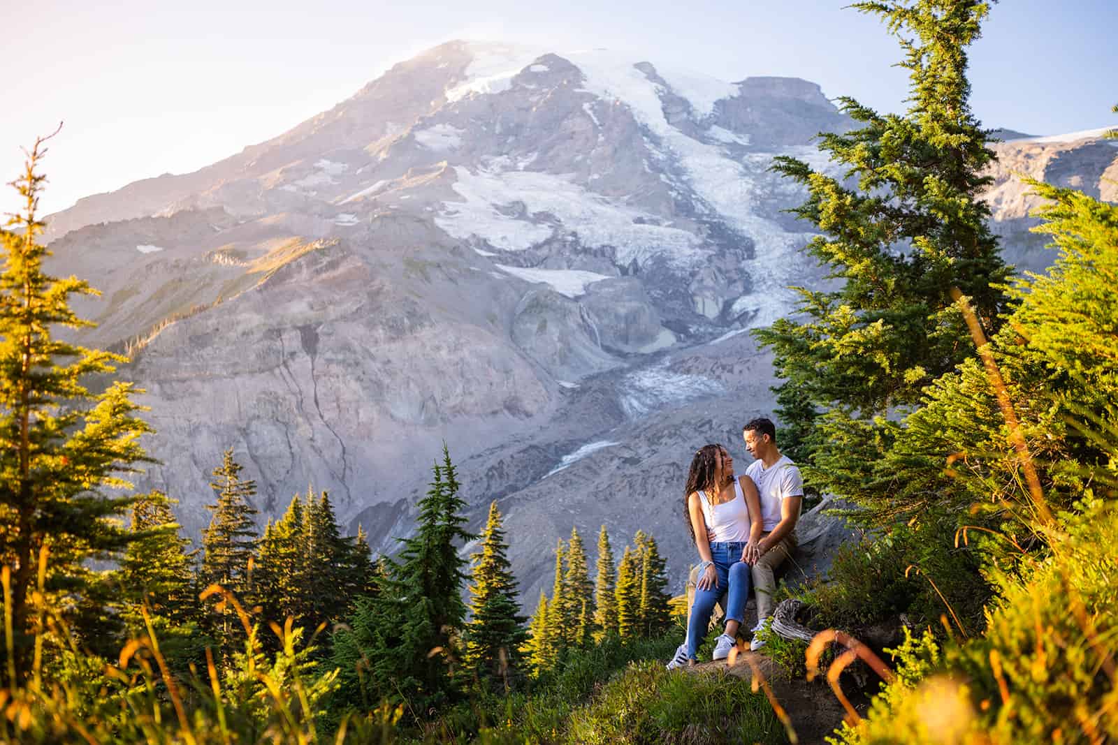 A couple cuddles in the shade of a pine tree in front of a majestic mountain backdrop during their fall engagement photos at Mount Rainier National Park.