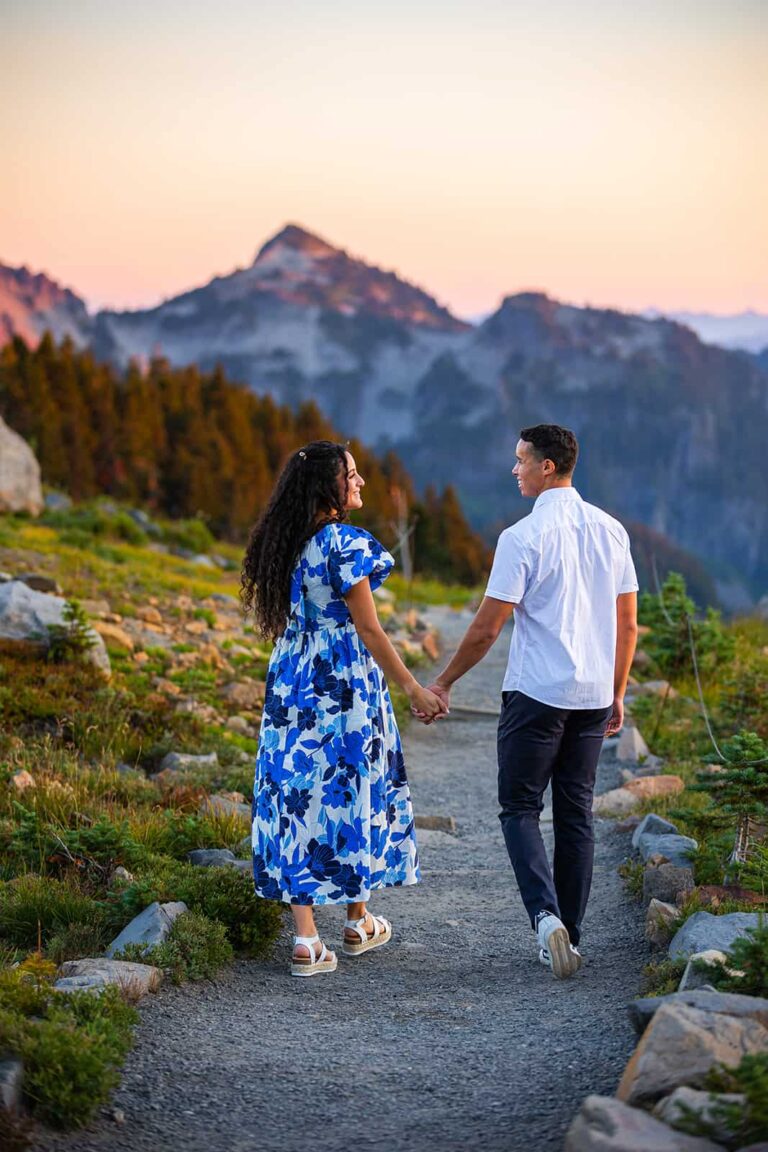 A couple holds hands and smiles at one another as they walk towards mountains glowing pink in the distance.