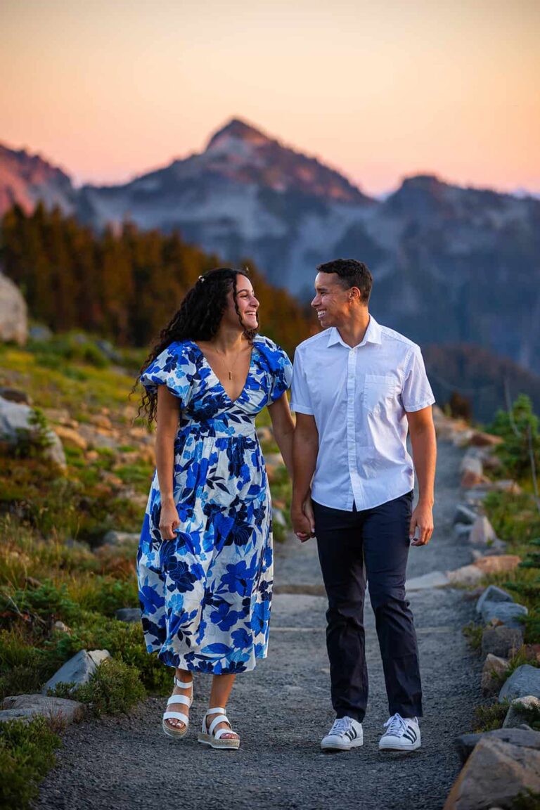 A woman in a blue floral dress and a man in a white shirt walk along a gravel path during their engagement session at Mount Rainier National Park in September.