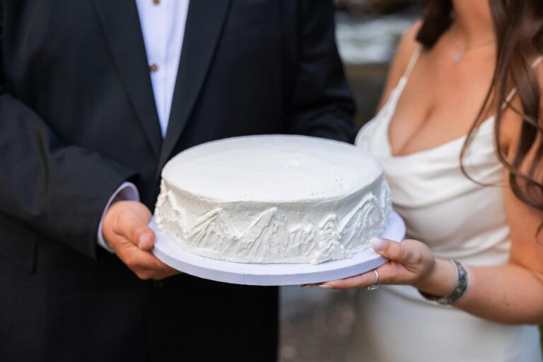 A bride and groom hold a white wedding cake decorated with simple mountains and pine trees.