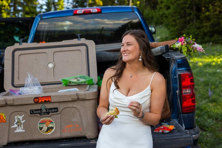 A bride smiles while eating her sandwich in front of a cooler sitting in the tailgate of a pickup truck during her adventure elopement in Wyoming.