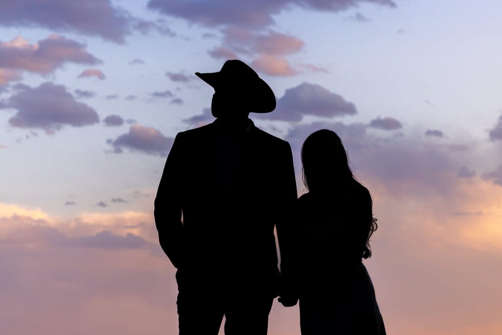 A silhouette of a man and woman standing side by side against a colorful sunset. The man is wearing a cowboy hat.