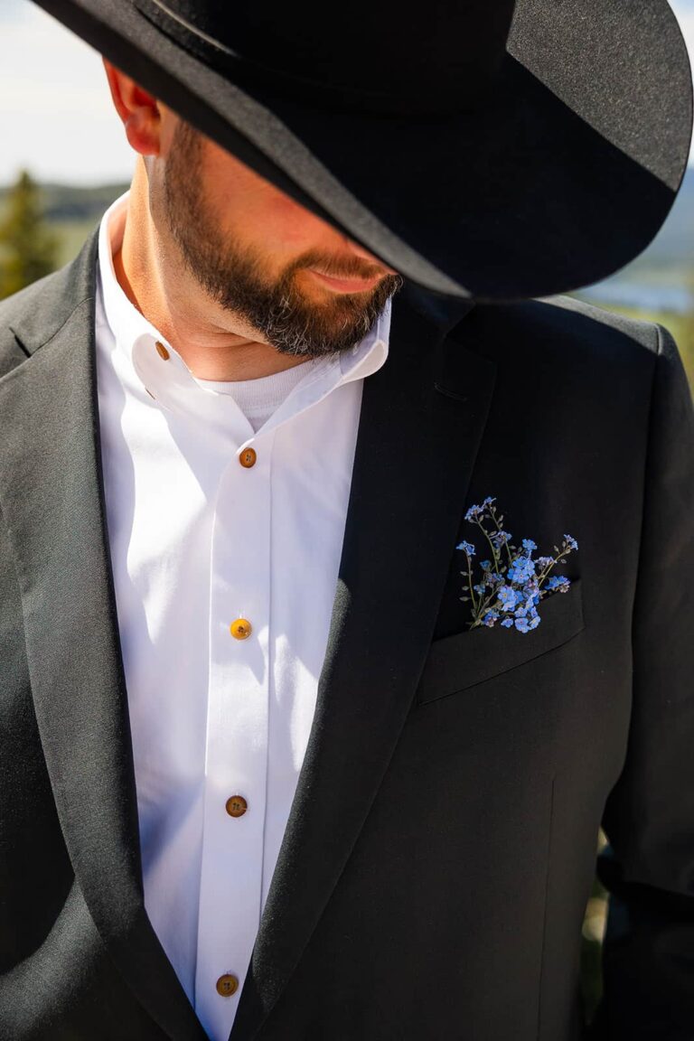 A groom wearing a black cowboy hat with a small boutonniere of blue flowers in his pocket poses for a picture during his western-inspired elopement in Wyoming.