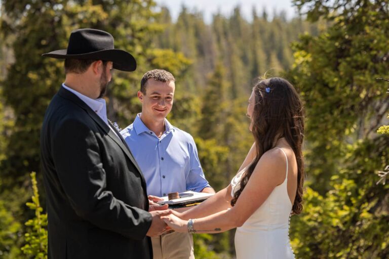 A bride and groom stand in front of one of their friends who is officiating their meadowlark lake elopement in the forests of Wyoming.
