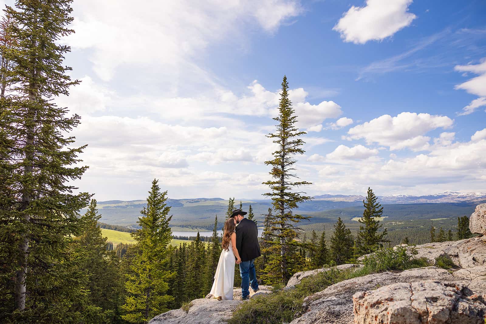 A newly married couple kissing in their wedding cloths on a cliff surrounded by pine trees during their Meadowlark Lake wedding in Wyoming.