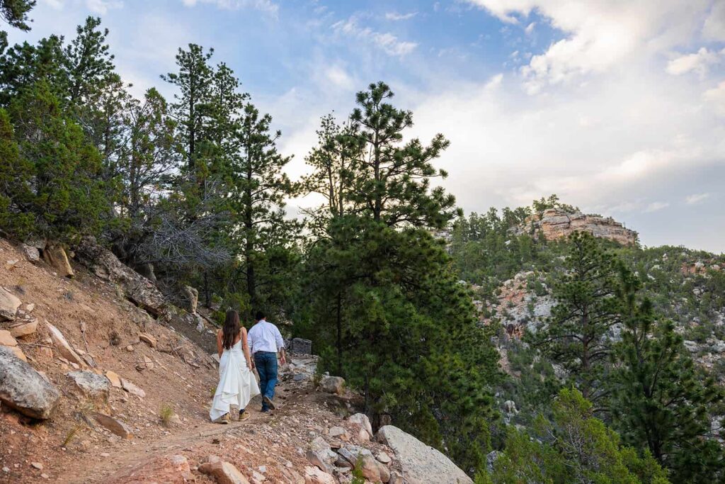 A couple hike up a trail in their wedding clothes surrounded by pine trees.