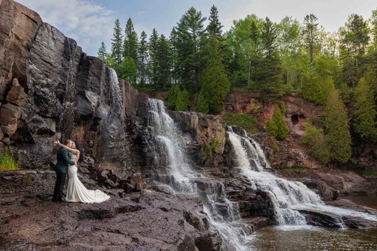A couple embrace in front of Gooseberry Falls during their Minnesota elopement with Forever and Evergreens.