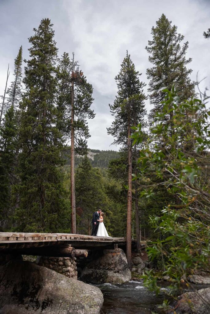 A bride and groom stand on a wooden bridge among towering pines during their elopement with Forever and Evergreens.