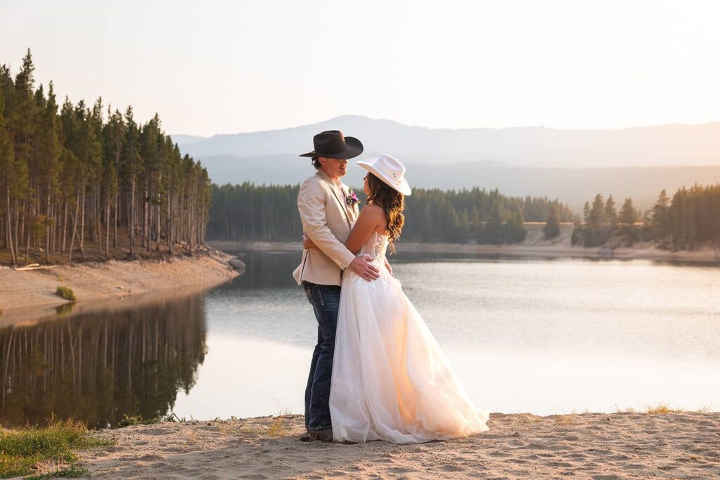 A bride and groom wearing cowboy hats embrace in front of a lake during their adventure elopement with MN elopement photographers Forever and Evergreens.