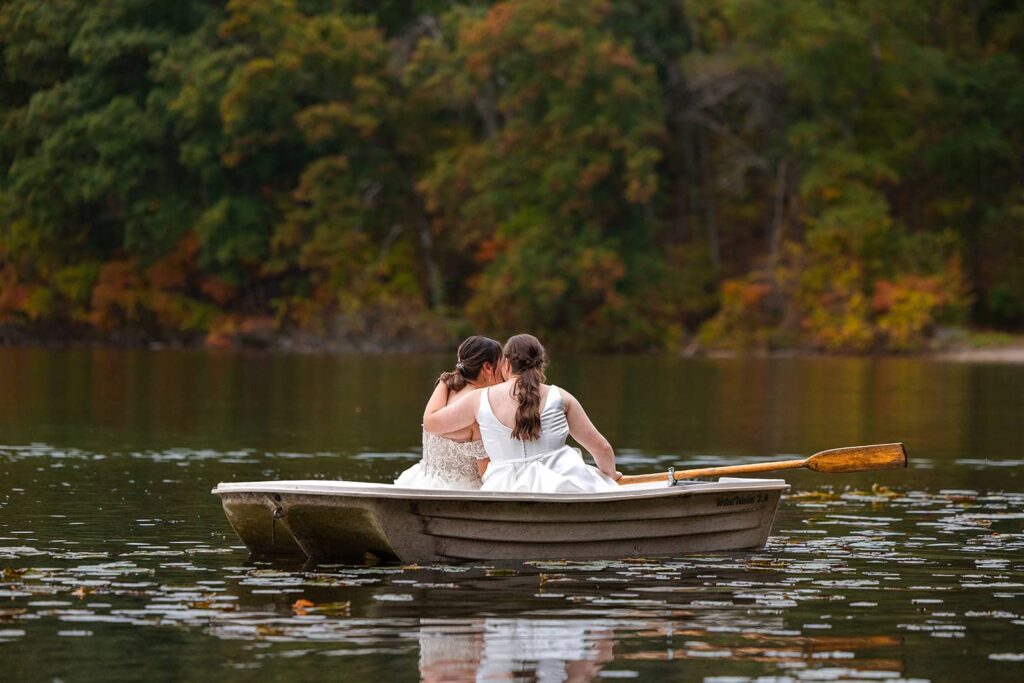 Two brides embrace while rowing a boat in a lake during their summer camp wedding.