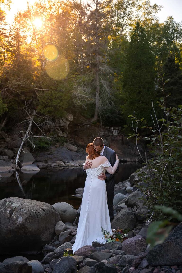 A couple embrace on the bank of a river during their Midwest elopement with Minnesota elopement photographers, Forever and Evergreens.