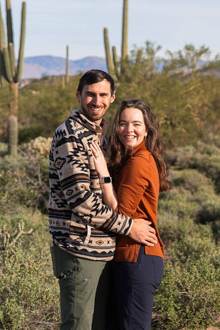 Tali and Dom, the photographers behind Forever and Evergreens Elopement Photography, embrace and smile at the camera while standing among cacti in the desert.